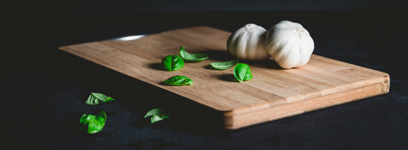 Photographie sur fond noir d'une planche à découper en bois avec une gousse d'ail et quelques feuilles de basilic