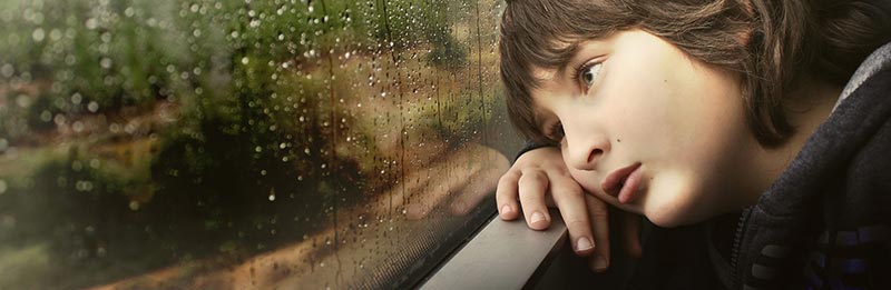 Photographie d'un enfant fatigué, la tête posée sur ses mains, regardant la pluie tomber à travers la fenêtre d'un train, pour illustrer les carences en fer et la fatigue de l'anémie suite au manque d'aliments riches en fer.