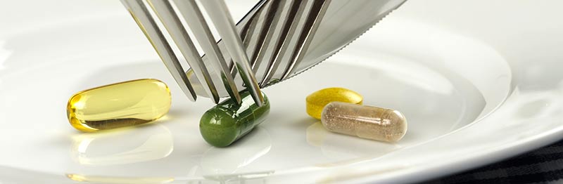 Photographie couleur de 4 pilules de compléments alimentaires de couleur jaune, transparente, beige et verte, posées dans une assiette blanche, avec quelqu'un essayant de les couper pour les manger à l'aide d'une fourchette et d'un couteau. Les compléments alimentaires sont-ils dangereux ? Voici quelques éléments de réponse avec le laboratoire Shavi.