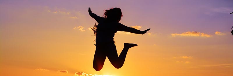 Photographie en couleur d'une femme à contre-jour sautant de joie devant un magnifique couché de soleil coloré