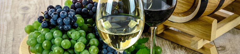 Photographie d'un verre de vin rouge et d'un verre de vin blanc devant une grappe de raisins rouges et blancs et un petit tonneau en bois, pour illustrer le fait que le vin est une boisson riche en fer.