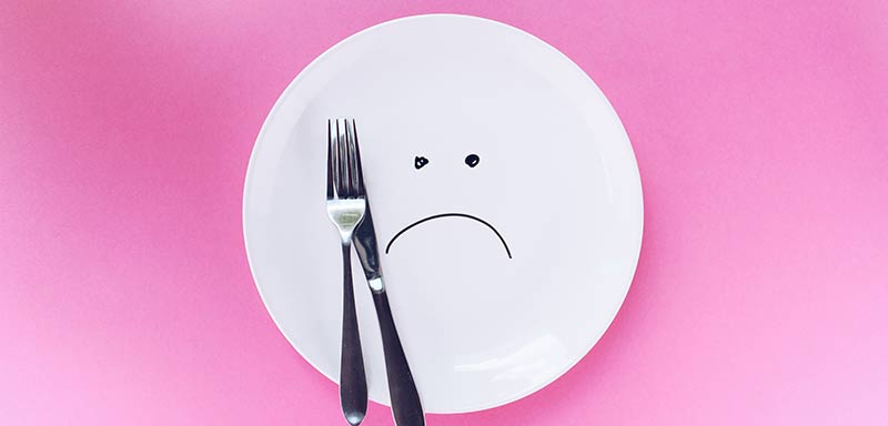 Photographie d'une assiette blanche vide avec un smiley triste et une paire de couverts sur un fond rose afin d'illustrer les dangers du jeûne et des régimes amaigrissants restrictifs pour perdre du poids.