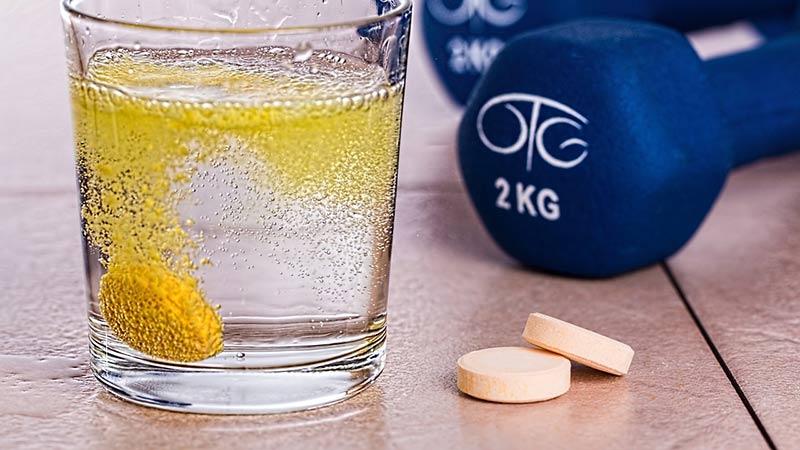 Comprimé de vitamine C effervescent dans un verre d'eau sur une table avec deux haltères bleues pour femme pour démontrer les dangers des compléments alimentaires pour perdre du poids.