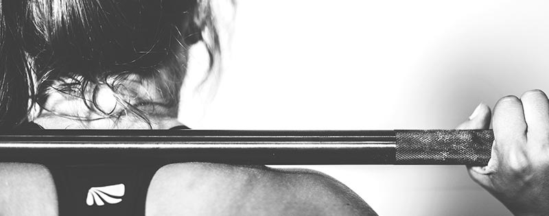 Photographie en noir et blanc du dos et de la nuque d'une femme en tenue de sport en train de lever une barre de musculation pour perdre du poids