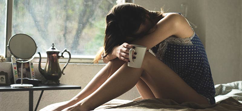 Photographie d'une femme triste, la tête entre ses mains, assise sur son lit avec une tasse de thé à la main car elle n'arrive pas à perdre du poids