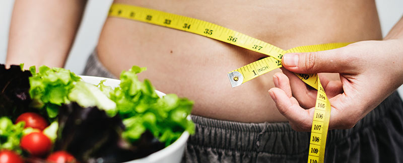 Photographie en gros plan d'une femme avec un peu de graisse abdominale en train de mesurer son tour de taille avec un mètre ruban et tenant une salade dans sa main droite pour perdre du poids