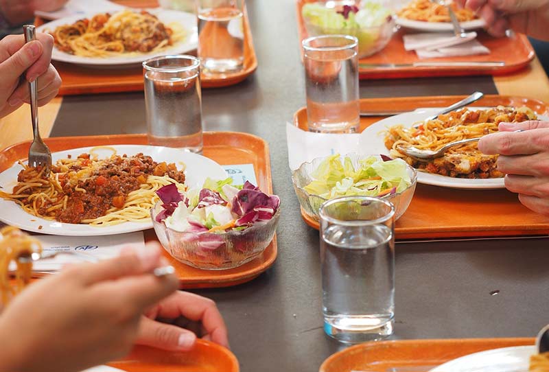 Photographie d'une table avec plusieurs plateaux oranges de self contenant des assiettes de spaghettis bolognaise, une salade vert et un vert d'eau. Les convives sont en train de manger un repas de midi typique des cantines scolaires et de la restauration collective.