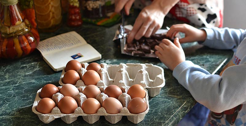 Importance de l'éducation alimentaire : maman en train de cuisiner avec son enfant un gâteau au chocolat. Photogrpahie en gros plan des mains des membres de la famille qui participent à cet atelier cuisine. Des œufs sont posés sur une table en marbre et on aperçois également une tablette de chocolat et un livre de cuisine ouvert.