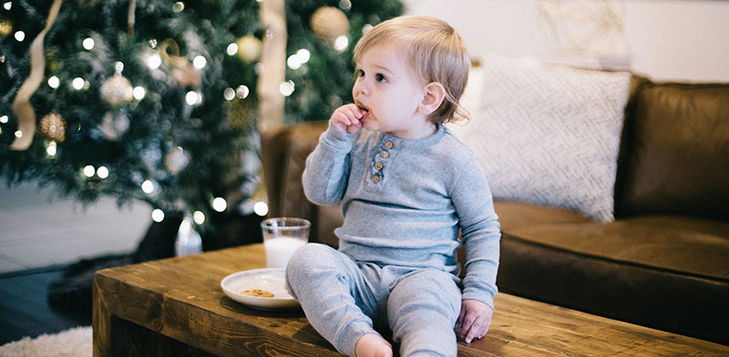 Enfant en pyjama gris, assis sur une table basse en bois devant un sapin de Noël illuminé, en train de prendre son goûter : des gâteaux sucrés et un verre de lait. La santé des enfants passera par l'éducation alimentaire.
