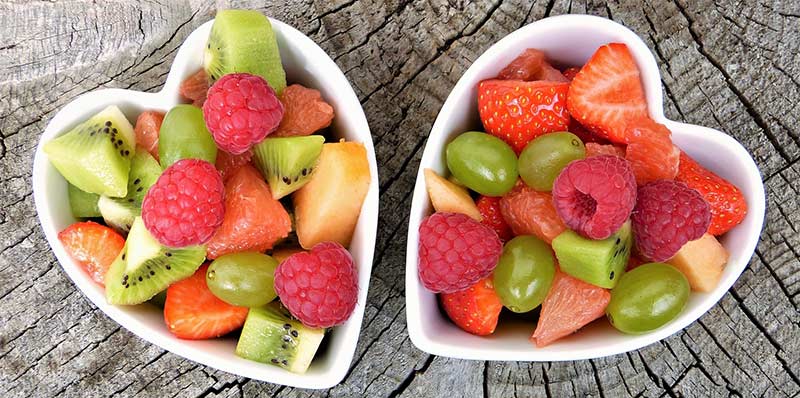 Photographie couleur de deux bols blancs en forme de cœur contenant des fruits frais bourrés de vitamine, posés sur une table en bois brut.