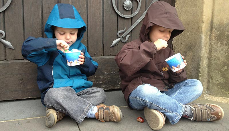 Photographie de deus enfants assis en tailleur dans la rue devant une porte en bois, habillés de jeans et d'anorak à capuche, en train de manger une glace dans un petit pot bleu.