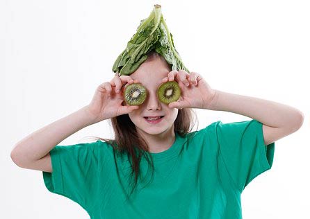 Manipulation de légumes par les enfants : Photographie d'une fille avec un tshirt vert qui a placé des feuilles de chou sur sa tête et deux tranches de kiwi sur ses yeux.