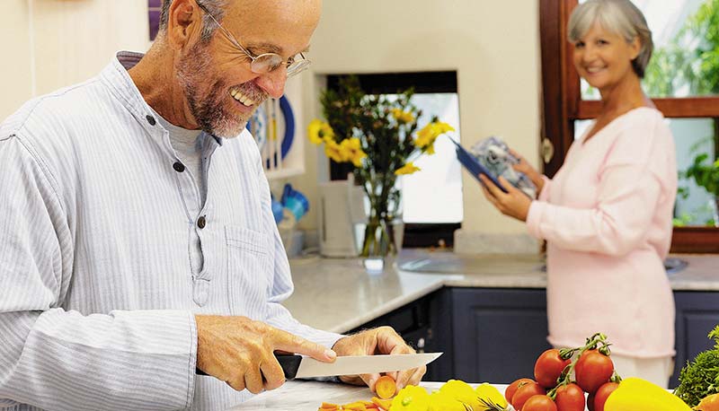 Photographie d'un homme cuisinant un régime alimentaire adapté, spécial contre l'arthrose. Il découpe des légumes crus et colorés sur une planche, pendant que sa femme retraitée le regarde. Mangez mieux, bougez plus pour dire Stop Arthrose !