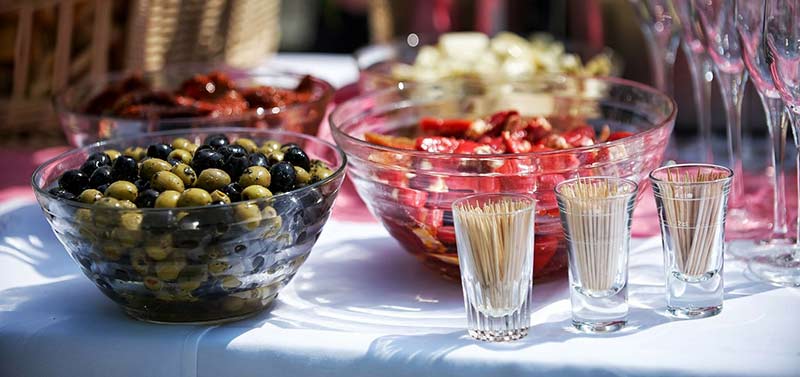 Photographie couleur d'un buffet apéritif posé en extérieur, contenant un saladier d'olives vertes et noires et différents légumes marinés, ainsi que des verres à champagne et des cure-dents.