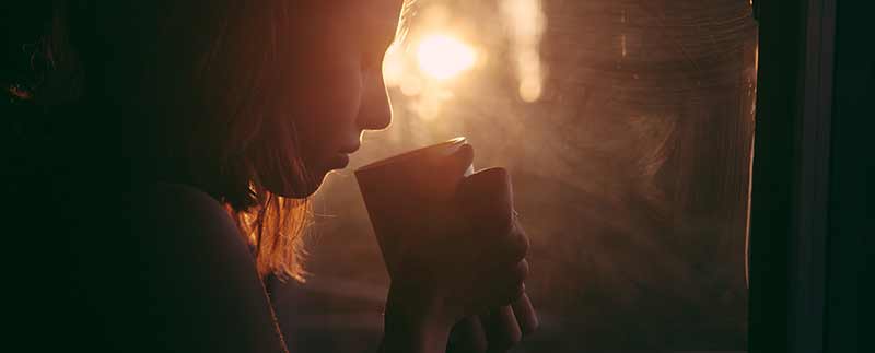 Femme en contre-jour buvant un thé chaud devant sa fenêtre : attention aux risques de carences en fer.