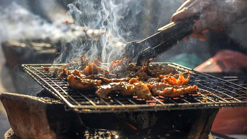 Photographie couleur d'une personne en train de faire griller de la viande sur un barbecue, ce qui génère de la fumée.