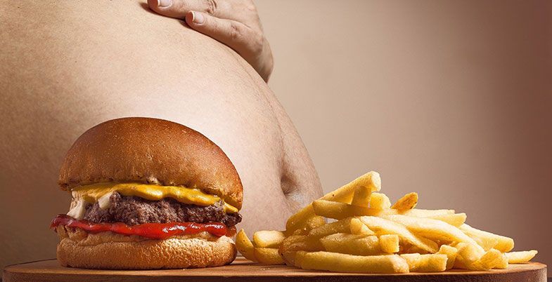Diabète : photographie du ventre gonflé d'une personne obèse devant un hamburger et des frites, illustrant les problèmes de surpoids liés à la malbouffe, qui entrainent une explosion du diabète.
